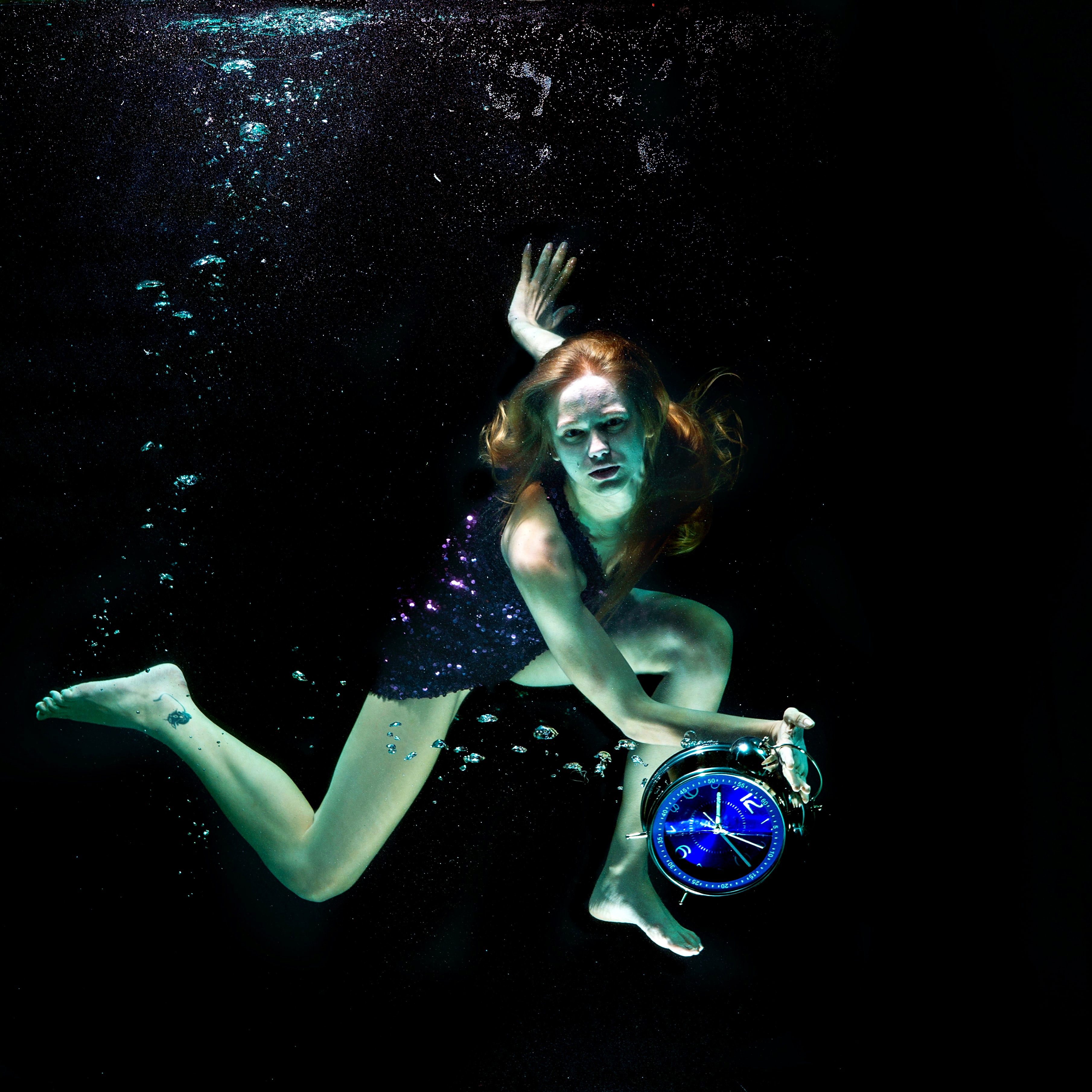 Frau mit violettem Paillettenkleid hält unter Wasser große Uhr ins Bild